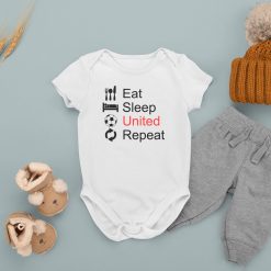 Flat lay of a white baby onesie with the phrase "Eat Sleep United Repeat" in black and red text with icons for eating, sleeping, a soccer ball, and a repeat symbol, displayed with cozy winter baby accessories including a knit hat, gray pants, and fur-lined boots; Manchester United themed baby clothing for football fans.