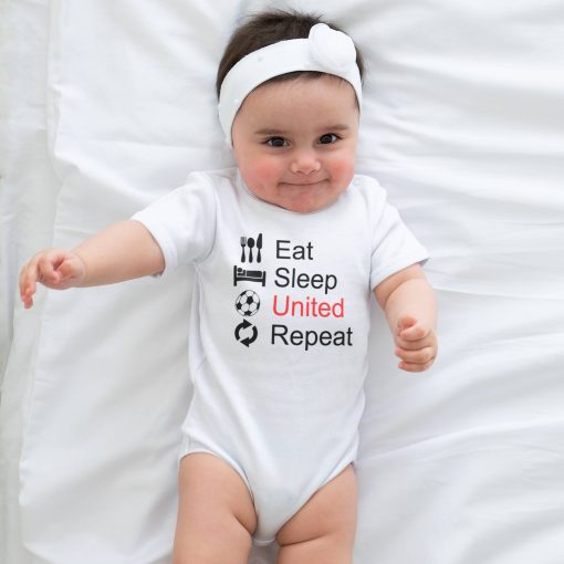 Eat Sleep United 03 Smiling baby girl lying on a white bed wearing a white headband and a white baby onesie printed with black and red text reading "Eat Sleep United Repeat" accompanied by icons for utensils, a bed, a soccer ball, and a repeat symbol; humorous Manchester United football-themed infant outfit.