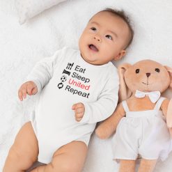 Eat Sleep United 05 Baby boy in a long-sleeve white onesie lying on a white fluffy blanket beside a beige plush bunny, wearing clothing that reads "Eat Sleep United Repeat" with black and red text and soccer icons; cozy and cute Manchester United baby gift idea.