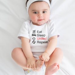 Eat Sleep United 07 Baby girl sitting on a white sheet, holding her feet, wearing a white onesie with "Eat Sleep United Repeat" slogan in black and red with football-related graphics; funny and stylish baby bodysuit for Manchester United supporters.
