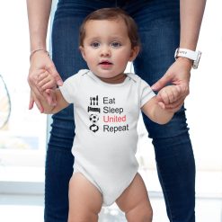Toddler girl supported by an adult, wearing a white baby bodysuit that reads "Eat Sleep United Repeat" in black and red with icons of utensils, bed, soccer ball, and arrows; sporty Manchester United themed baby fashion.