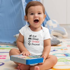 Happy baby boy sitting indoors in a white short-sleeve onesie featuring the phrase "Eat Sleep United Repeat" with matching black icons and red “United” text, referencing Manchester United football club; playful soccer-themed baby apparel.