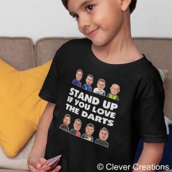 Young child wearing a black t-shirt featuring illustrated faces of the 2026 premier league darts players and the text "STAND UP IF YOU LOVE THE DARTS," ready for the excitement of the 2026 Premier League Darts.