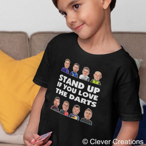 Young child wearing a black t-shirt featuring illustrated faces of the 2026 premier league darts players and the text "STAND UP IF YOU LOVE THE DARTS," ready for the excitement of the 2026 Premier League Darts.