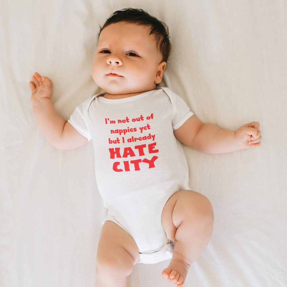 Baby lying on a white bed wearing a white onesie with red text that reads, 