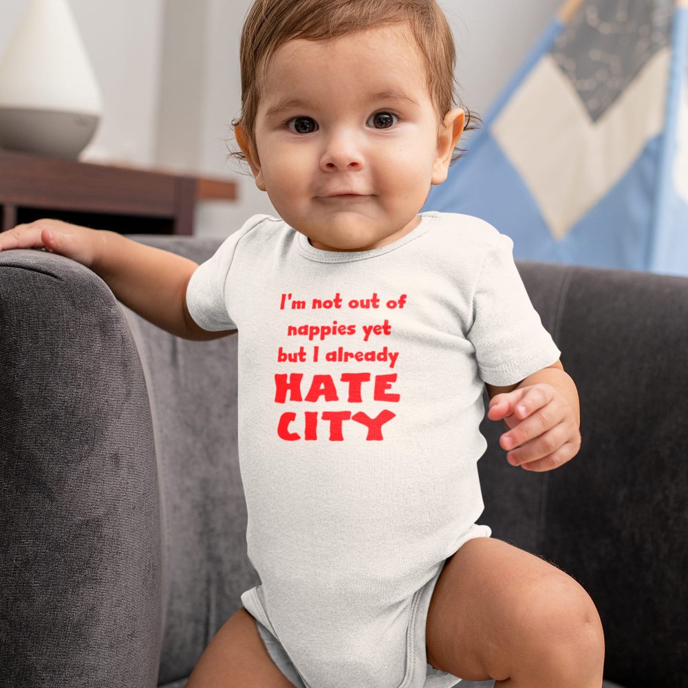 Baby standing on a dark gray couch wearing a white onesie with bold red text that reads, 