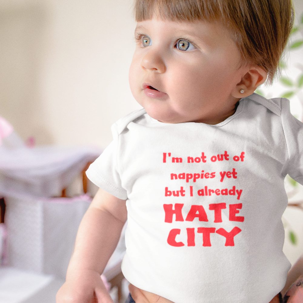 Close-up of a baby with light skin, light brown hair, and blue-green eyes, wearing a white onesie with bold red text that says, 