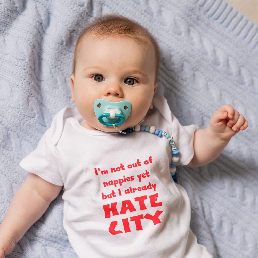 Baby lying on a light blue knitted blanket, wearing a white onesie with bold red text that reads, 