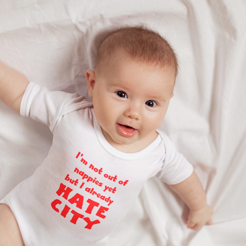 Smiling baby lying on a white sheet, wearing a white onesie with bold red text that reads, 