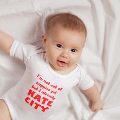Smiling baby lying on a white sheet, wearing a white onesie with bold red text that reads, "I'm not out of nappies yet but I already HATE CITY." The baby's playful expression and the humorous message on the outfit suggest early football rivalry, likely aimed at Manchester City FC. Ideal for SEO targeting keywords like “funny baby football onesie,” “anti-Man City baby clothes,” and “soccer rivalry infant apparel.”