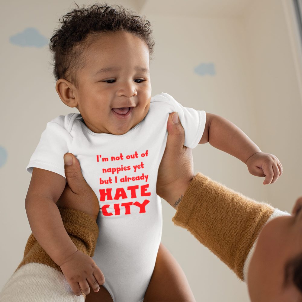 Smiling baby with curly hair held up by an adult, wearing a white onesie with bold red text that reads, 