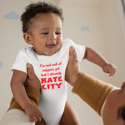 Hate-City-8 Smiling baby with curly hair held up by an adult, wearing a white onesie with bold red text that reads, "I'm not out of nappies yet but I already HATE CITY." The background features a softly lit nursery room with light-colored walls and cloud-shaped decals. The playful slogan on the outfit humorously expresses early football rivalry, likely referencing opposition to Manchester City FC, making the image ideal for sports fan baby apparel, football-themed merchandise, or humorous anti-Manchester City fan content.