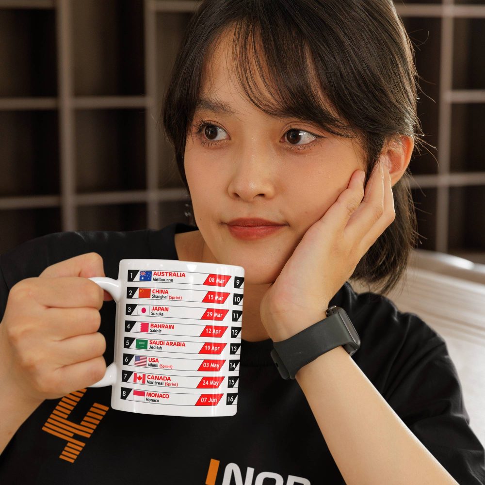 Woman in a Lando Norris shirt holding a white ceramic mug printed with the 2026 Formula One Grand Prix race schedule. Ideal F1 fan merchandise or motorsport-themed gift.