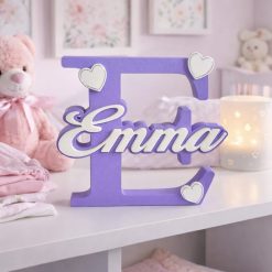 Personalised nursery letter name sign Emma on purple initial E with heart decorations, freestanding kids room décor displayed on nursery shelf with teddy bear and baby items. Available in multiple colour and adornment options.